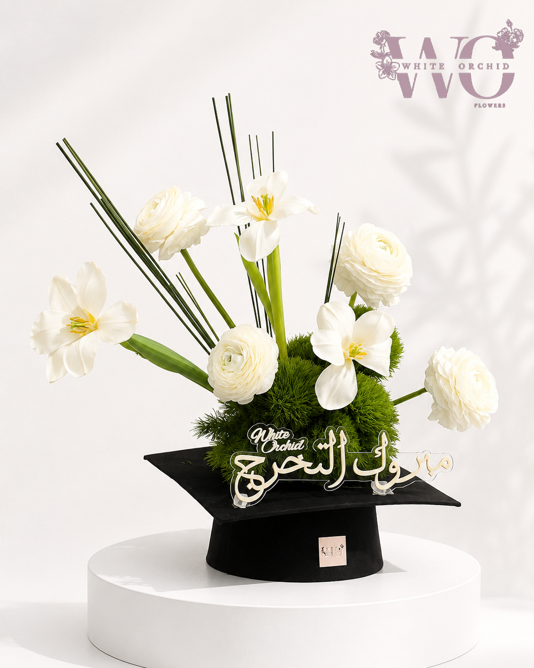Graduation flower arrangement with white flowers and greenery displayed in a graduation cap container.