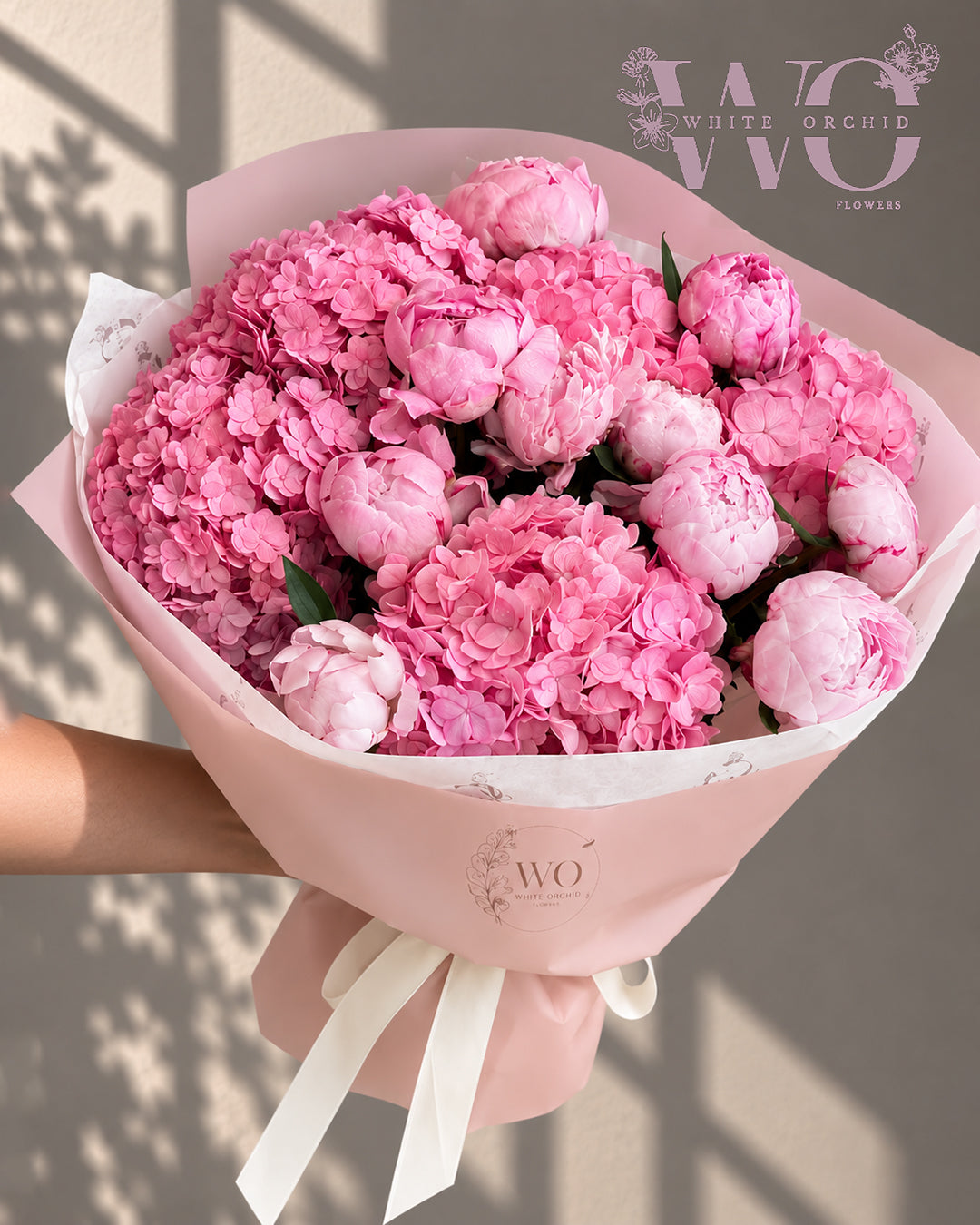 Pink peony and hydrangea hand bouquet wrapped in pink paper with white ribbon by White Orchid Flowers