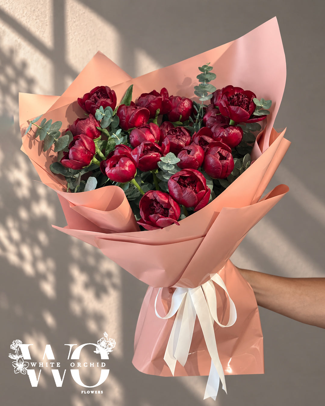 Red peony hand bouquet with eucalyptus, pink wrapping and white ribbon by White Orchid Flowers