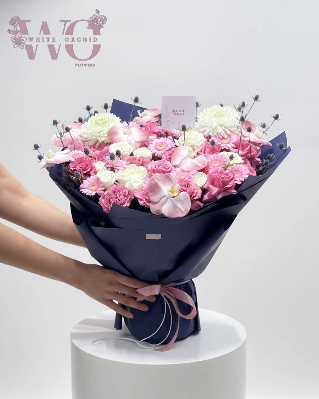 Luxury pink mixed flower bouquet with roses, orchids, and gerbera wrapped in navy paper with pink ribbon.