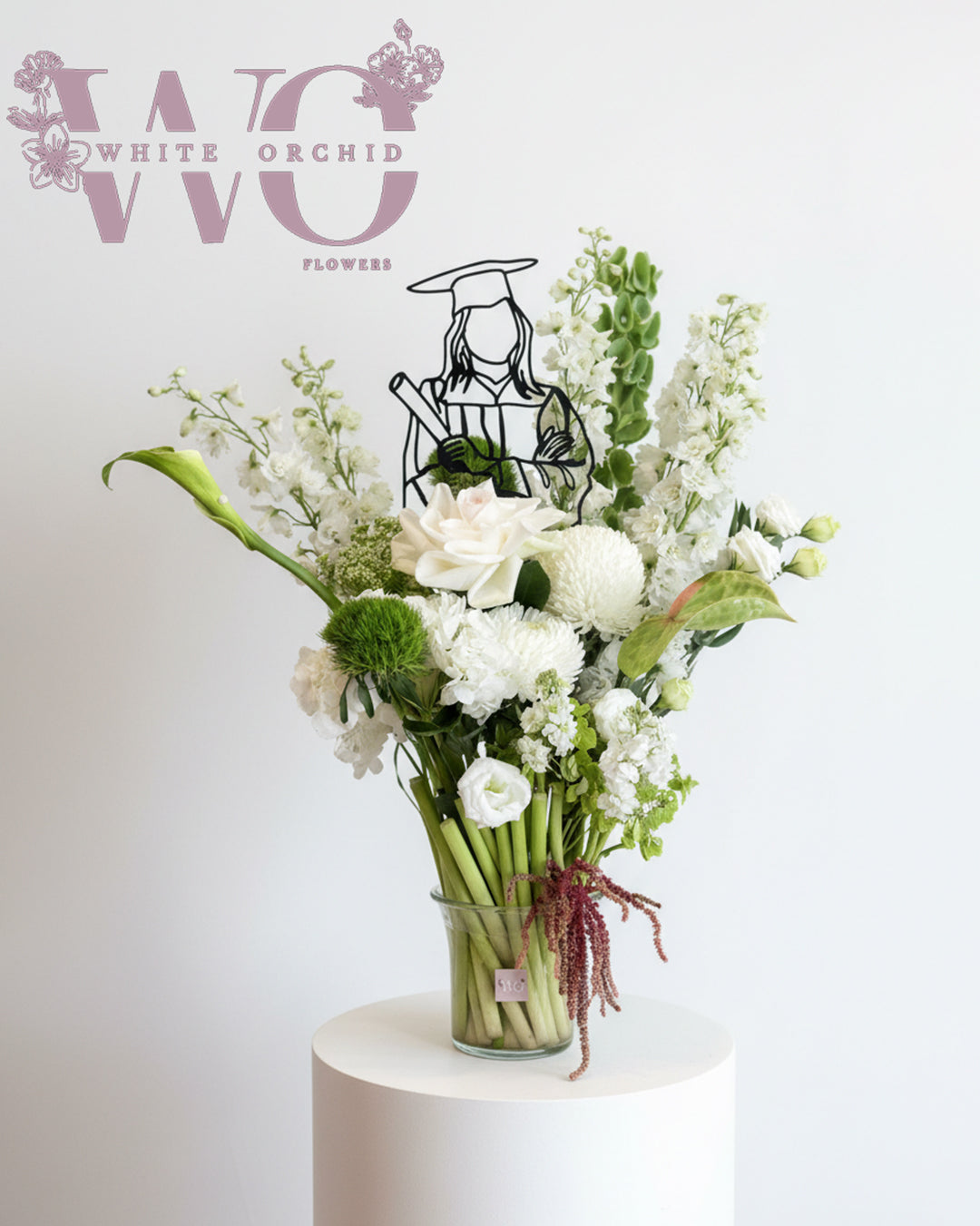 White graduation flower arrangement with roses, calla lily, and graduation topper in a glass vase.